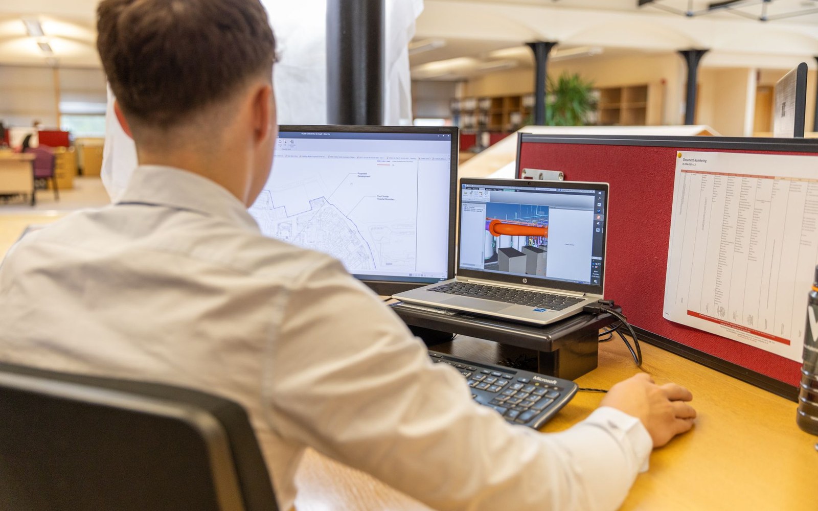 Man at a desk viewing site plans on a monitor and 3D model on a laptop