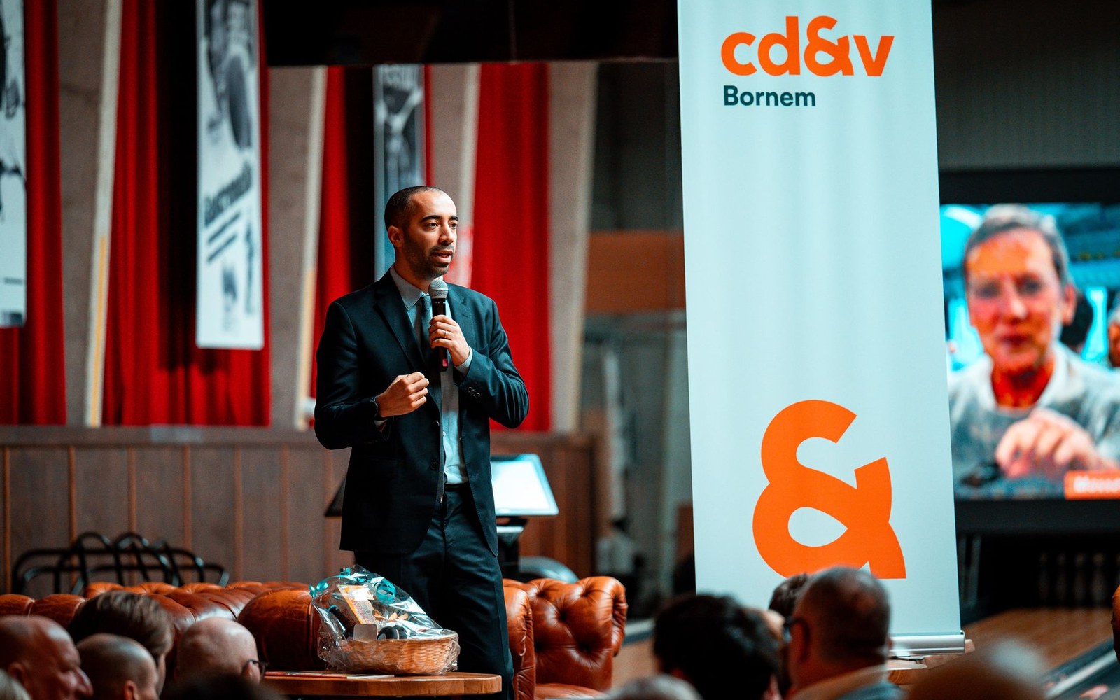 A man speaks into a microphone on stage beside a cd&v Bornem banner